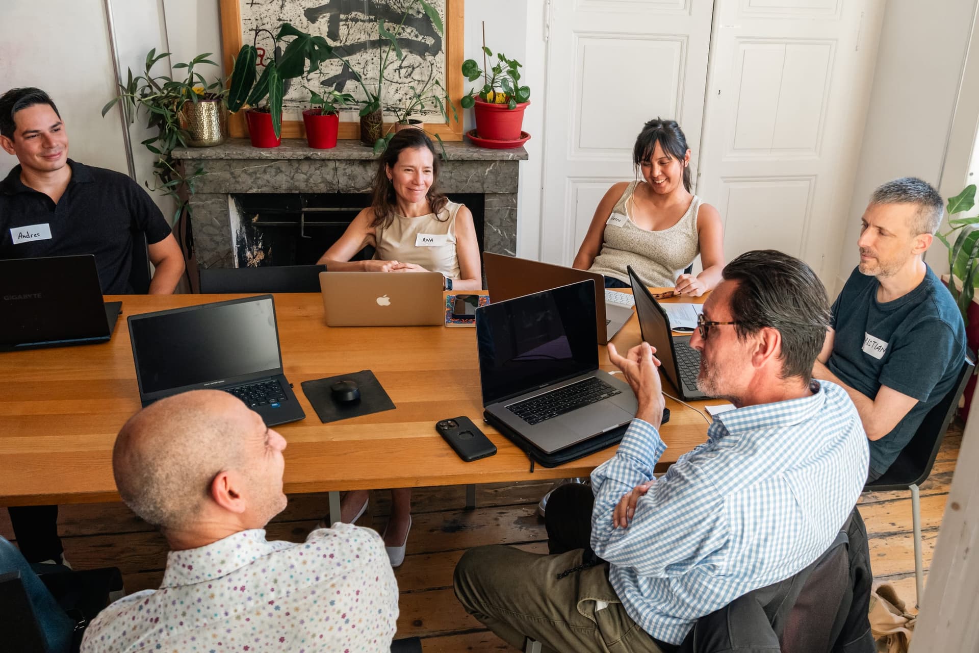 Workshop participants in discussion around a wooden table with laptops, beside a fireplace with plants