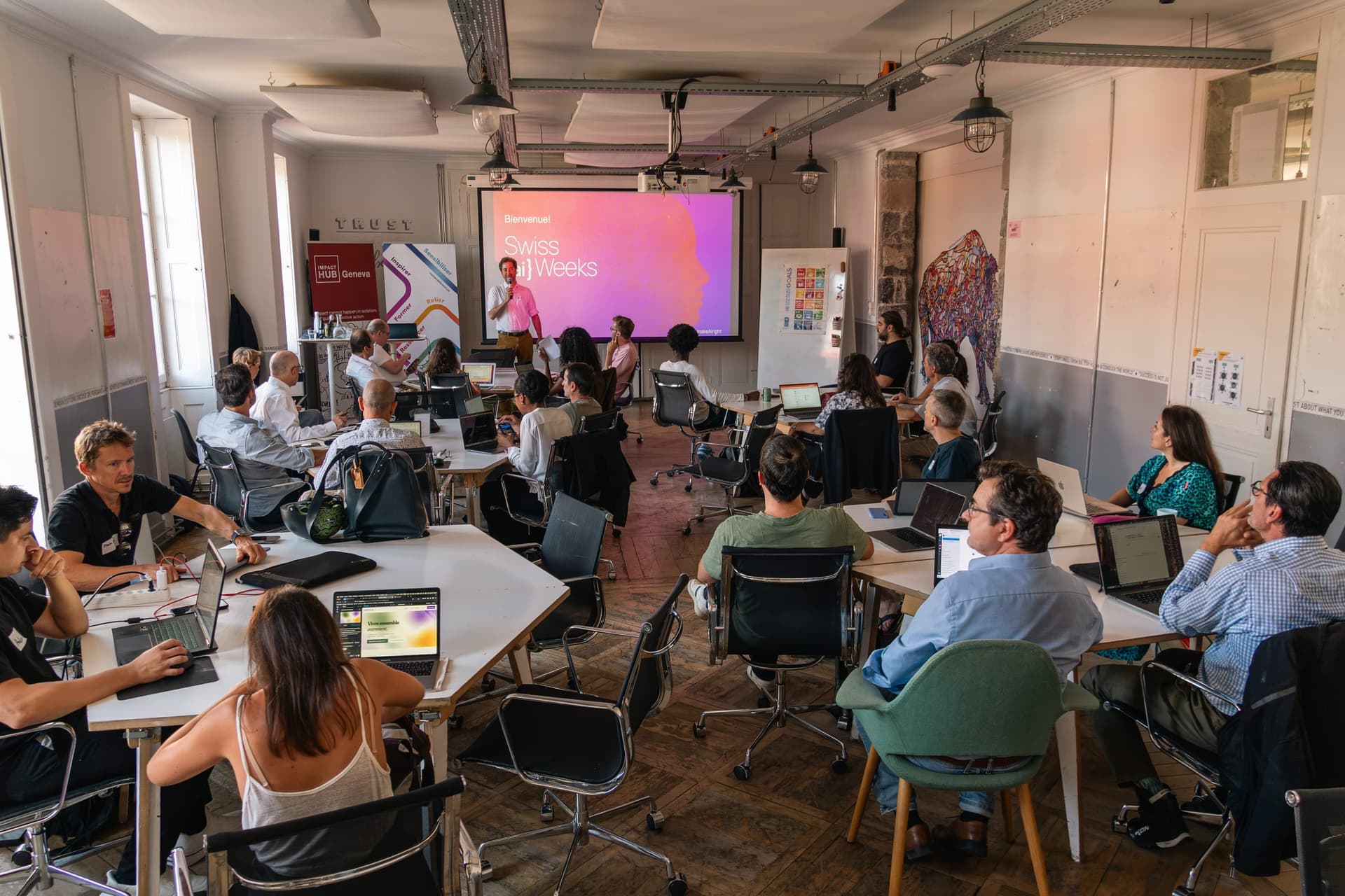 Attendees with open laptops facing a welcome slide for Swiss AI Weeks and a speaker with a microphone at Impact Hub Geneva