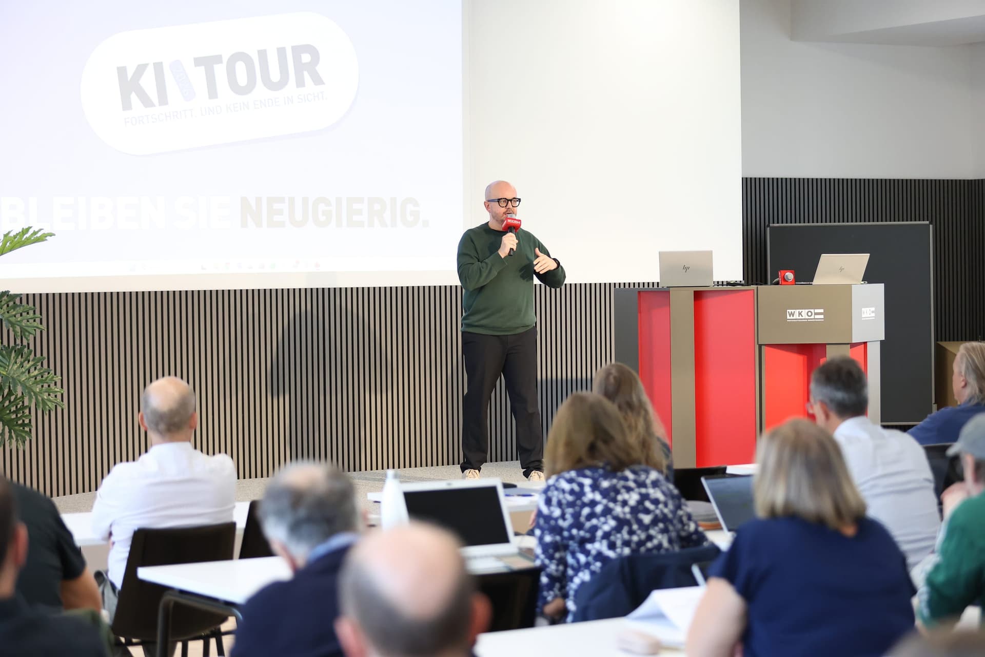 A presenter speaks below a KI TOUR slide while participants listen from their tables