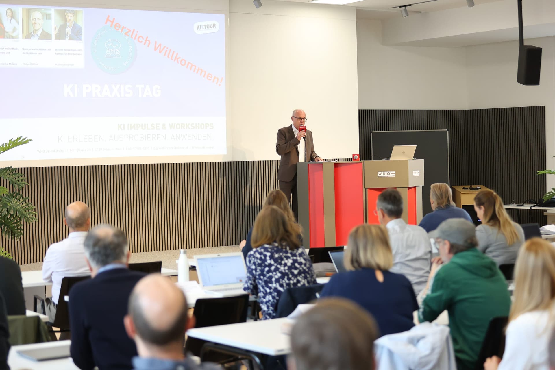 A speaker at a red WKO lectern beside a Herzlich Willkommen slide, with participants at white tables in the foreground