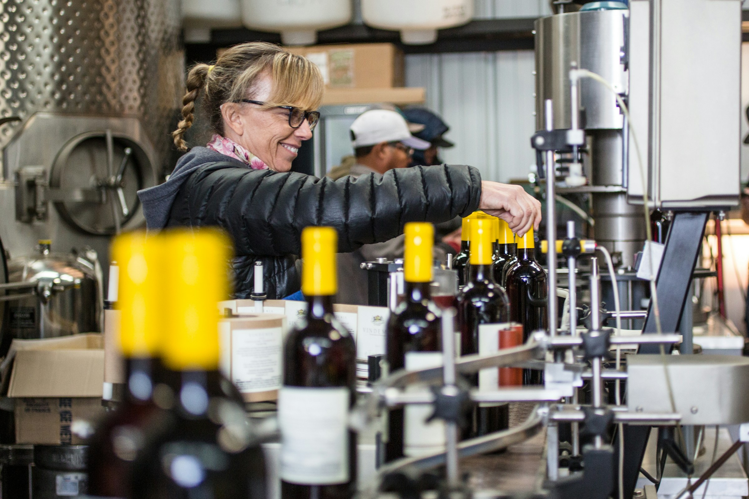 Operator guiding wine bottles along a bottling line beside stainless tanks