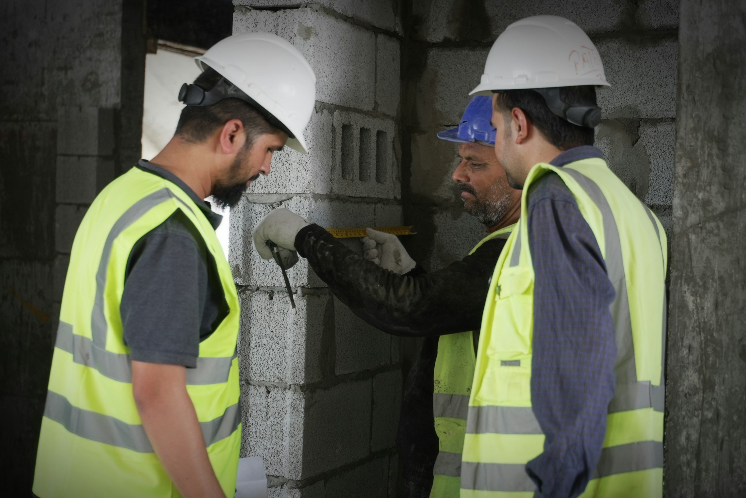 Three site workers in hard hats and vests measuring a block wall with a tape measure